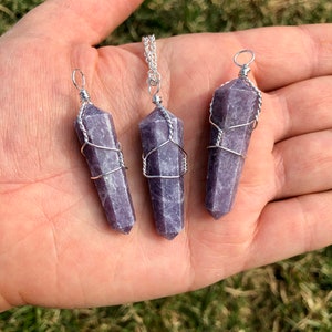 May include: Three purple lepidolite crystal pendants wrapped in silver wire. The pendants are of different sizes and are hanging from a silver chain.