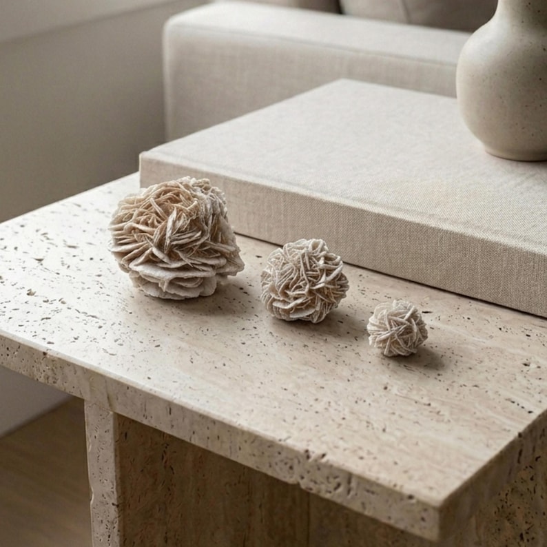 May include: Three desert rose formations in varying sizes sit on a light-colored stone table. The formations have a layered, textured appearance with a beige color. A beige book and vase are also on the table, creating a neutral-toned, natural aesthetic.