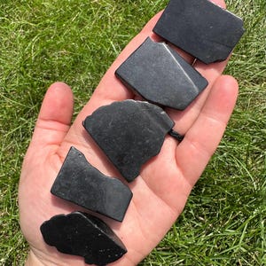 May include: Five black obsidian stones of various shapes and sizes. The stones are smooth and polished, with a deep black color.