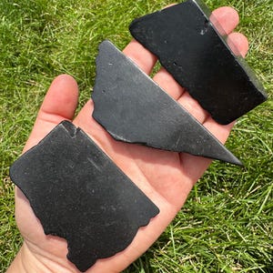 May include: Three black obsidian stones, each with a unique shape and smooth, polished surface. The stones are held in a hand, showcasing their size and texture.