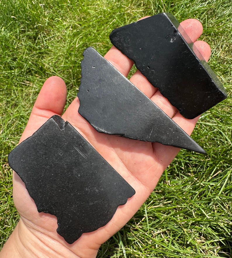 May include: Three black obsidian stones, each with a unique shape and smooth, polished surface. The stones are held in a hand, showcasing their size and texture.