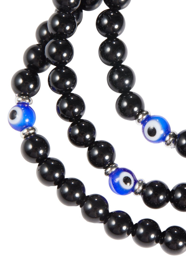 May include: Close-up of three strands of black beaded bracelets. Each strand features large, glossy black beads. Interspersed are blue and white evil eye beads, adding a pop of color. The bracelets are strung on a silver-toned metal.
