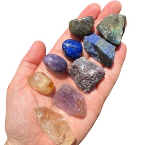 May include: A collection of eight tumbled stones in various colors, including amethyst, citrine, labradorite, and lapis lazuli. The stones are arranged in a hand, with the largest stone at the top and the smallest stone at the bottom.