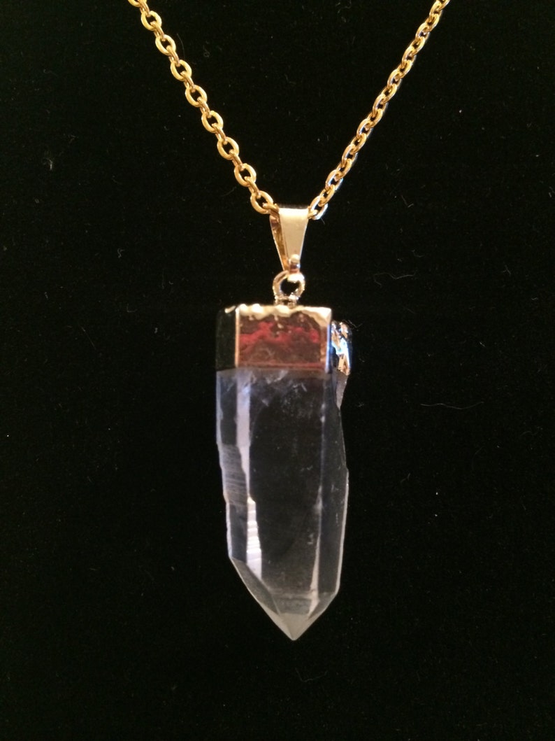 Puede incluir: Un collar de cadena dorada con un colgante de cristal de cuarzo transparente. El colgante est&aacute; facetado y tiene una tapa dorada en la parte superior.