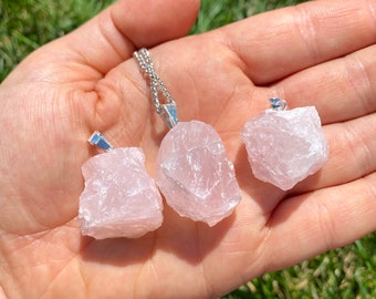 May include: Three raw rose quartz pendants on silver chains. The pendants are different sizes and shapes, with a natural, rough texture. The quartz is a pale pink color.