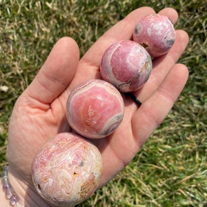 May include: Four pink and white striped spheres of various sizes, made of polished pink calcite, are held in a hand. The spheres are smooth and have a natural, organic look.