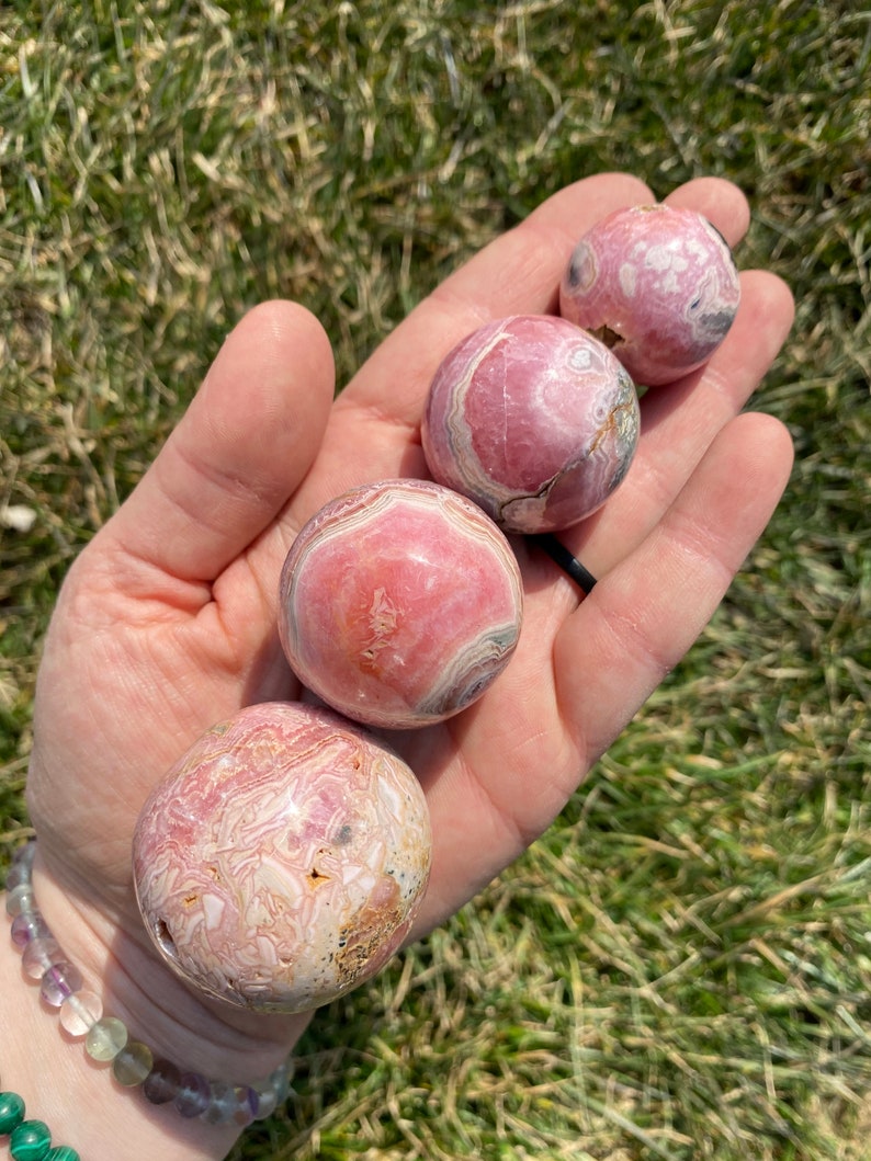 May include: Four pink and white striped spheres of various sizes, made of polished pink calcite, are held in a hand. The spheres are smooth and have a natural, organic look.