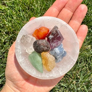May include: A white selenite bowl filled with seven small, colourful crystals. The crystals are red, orange, yellow, green, blue, purple, and clear.