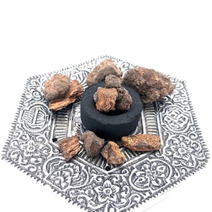 May include: A silver hexagonal incense burner with intricate designs, featuring a black charcoal disc with brown resin pieces on top.