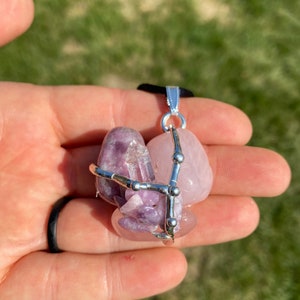 May include: A silver wire cage pendant with three pink and purple crystals. The crystals are held together by a silver wire cage with small silver beads. The pendant is hanging from a black cord.