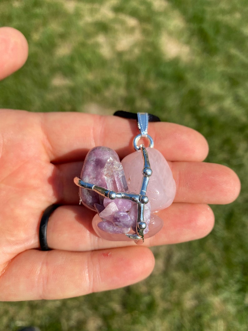May include: A silver wire cage pendant with three pink and purple crystals. The crystals are held together by a silver wire cage with small silver beads. The pendant is hanging from a black cord.
