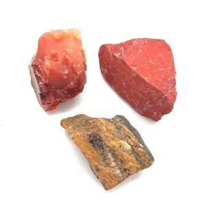 May include: Three rough, unpolished gemstone pieces. A red carnelian, a red jasper, and a tiger's eye.