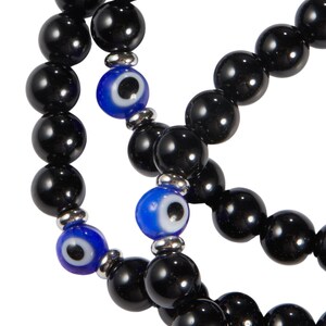 May include: Close-up of multiple strands of black beaded bracelets. Each strand features large, glossy black beads interspersed with blue and white evil eye beads. The bracelets are strung on a silver-colored metal.