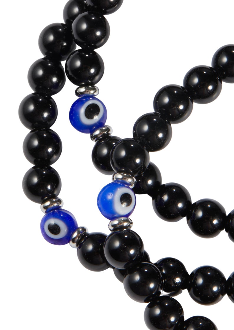 May include: Close-up of multiple strands of black beaded bracelets. Each strand features large, glossy black beads interspersed with blue and white evil eye beads. The bracelets are strung on a silver-colored metal.
