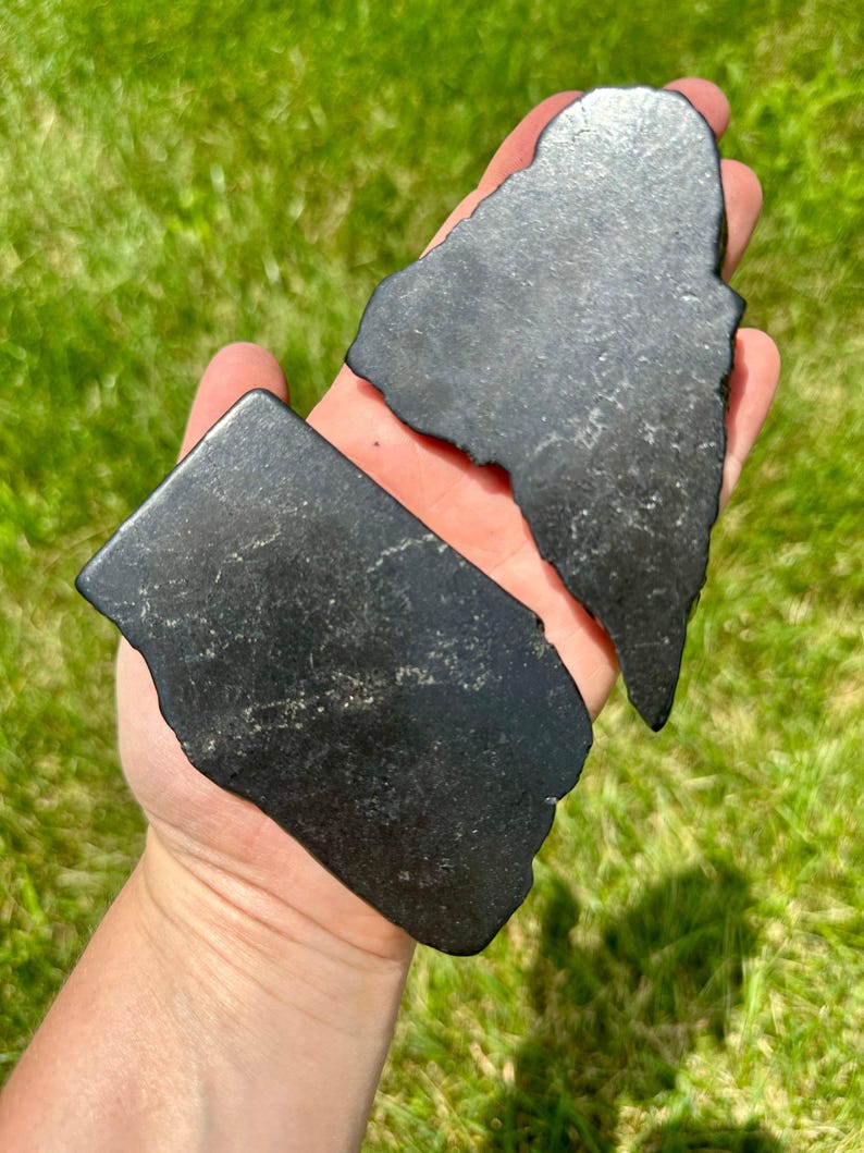 May include: Two pieces of black obsidian, one shaped like a triangle and the other a rectangle, are held in a hand.
