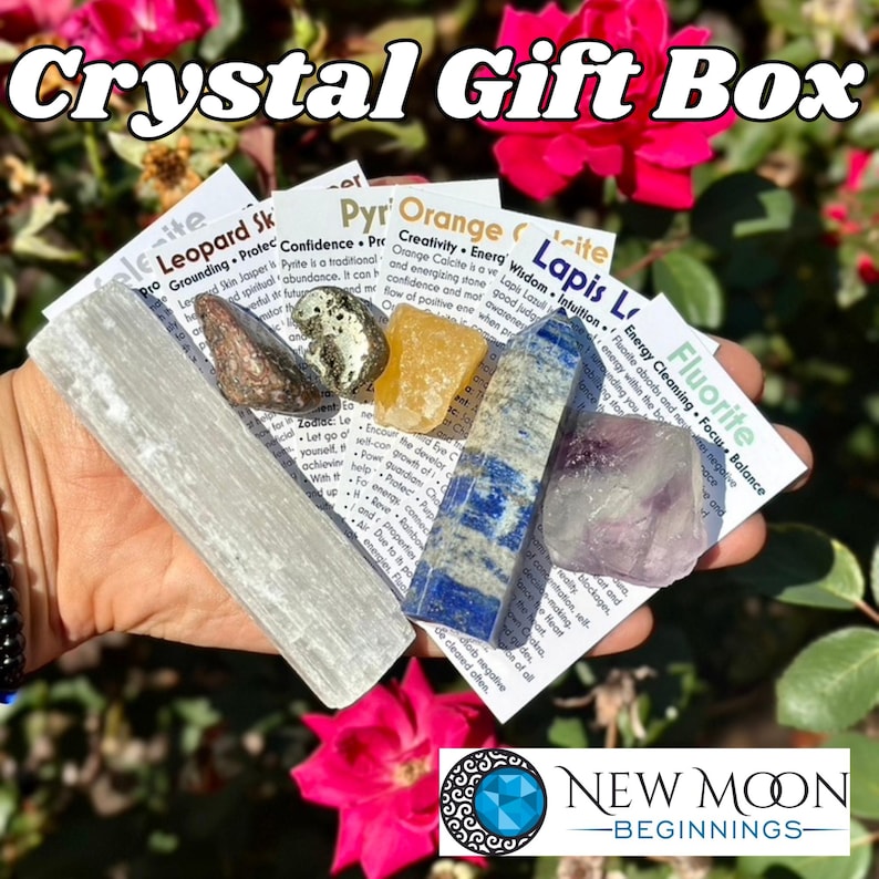 May include: A crystal gift box featuring a variety of stones, including selenite, jasper, pyrite, orange calcite, lapis lazuli, and fluorite. Each stone is accompanied by a card with information. The box is set against a backdrop of pink flowers.
