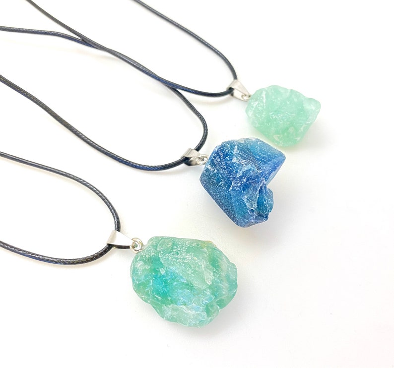 Puede incluir: Tres collares con colgantes de cristal de diferentes colores. Los collares son cordones negros con cierres plateados. Los colgantes son azul claro, azul oscuro y verde claro.