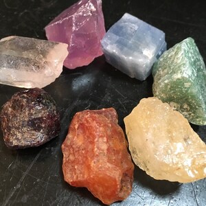 May include: Seven different coloured crystals arranged in a circle on a black surface. The crystals are clear, pink, blue, green, red, yellow, and brown.