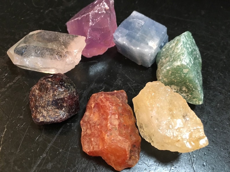 May include: Seven different coloured crystals arranged in a circle on a black surface. The crystals are clear, pink, blue, green, red, yellow, and brown.
