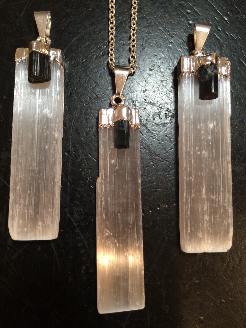 May include: Three selenite crystal pendants with black tourmaline accents. The pendants are on silver chains and have a polished, smooth finish.