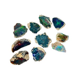 Puede incluir: Una colección de diez piedras de ópalo negro en bruto y natural con matices iridiscentes verdes, azules y rojos. Las piedras son de varios tamaños y formas, y algunas tienen una superficie rugosa y texturizada.