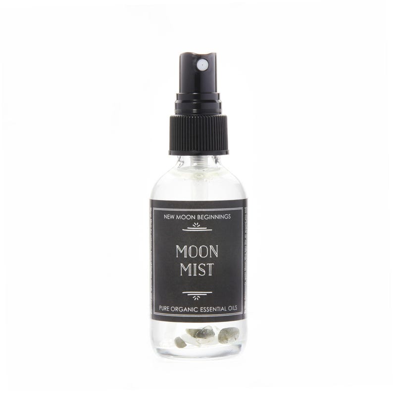 Puede incluir: Un frasco rociador de vidrio transparente con una tapa negra y una etiqueta negra. La etiqueta dice "MOON MIST" y "PURE ORGANIC ESSENTIAL OILS". La botella contiene peque&ntilde;as piedras transparentes. El texto "NEW MOON BEGINNINGS" est&aacute; en la parte superior de la etiqueta.