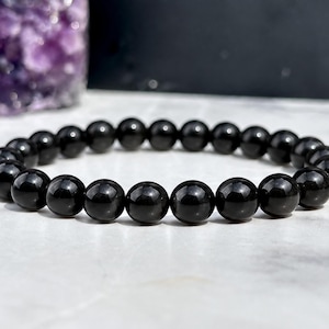May include: A stretch bracelet composed of numerous smooth, spherical black beads. The beads have a polished, reflective surface and are strung together in a continuous circular loop, creating a uniform and simple design for the wrist.