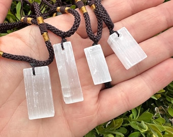 May include: Four rectangular selenite crystal pendants on brown braided cords. The cords have gold-colored accents. The pendants are held in a hand against a backdrop of green foliage. The selenite crystals are translucent and have a smooth surface.