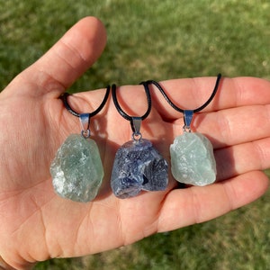 Puede incluir: Tres collares con colgantes de cristal en bruto. Los colgantes son verdes, azules y verdes. Los collares est&aacute;n en un cord&oacute;n negro con cierres plateados.