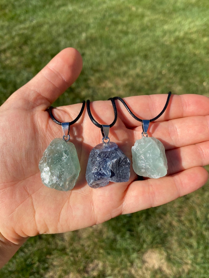 Puede incluir: Tres collares con colgantes de cristal en bruto. Los colgantes son verdes, azules y verdes. Los collares est&aacute;n en un cord&oacute;n negro con cierres plateados.