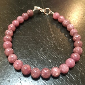 K&ouml;nnte beinhalten: Ein Armband aus rosafarbenem Lepidolith mit einem silbernen Verschluss. Das Armband besteht aus runden Perlen und ist etwa 18 cm lang.