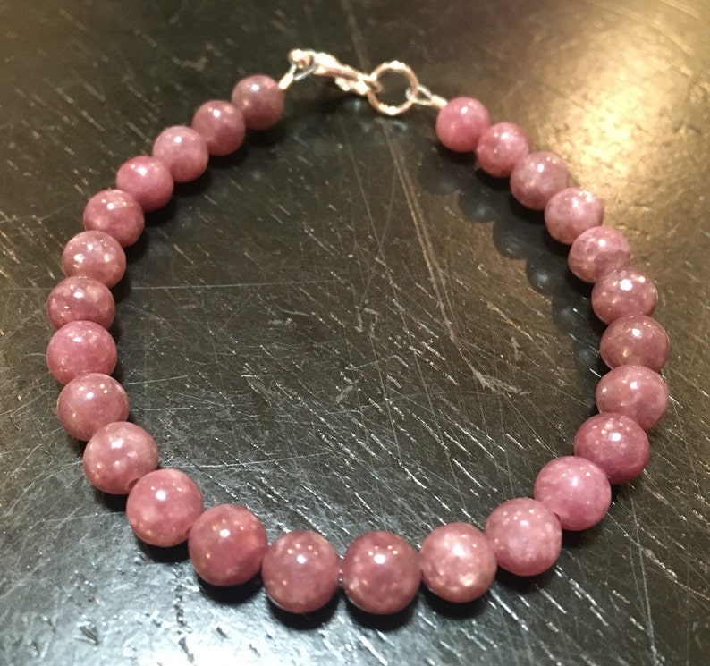 K&ouml;nnte beinhalten: Ein Armband aus rosafarbenem Lepidolith mit einem silbernen Verschluss. Das Armband besteht aus runden Perlen und ist etwa 18 cm lang.