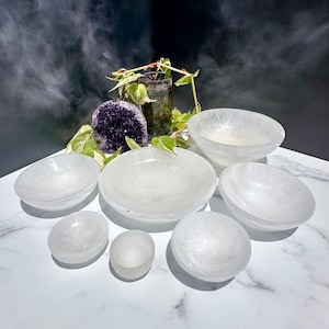 Pu&ograve; includere: Un set di sette ciotole di selenite bianca di diverse dimensioni, disposte su una superficie bianca. Le ciotole sono lisce e hanno una texture naturale e cristallina.
