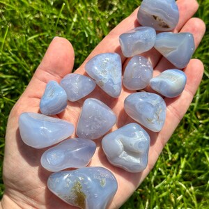 May include: A collection of smooth, tumbled blue lace agate stones. The stones vary in size and shape, with a pale blue color and white and brown inclusions. The stones are held in a hand against a backdrop of green grass.