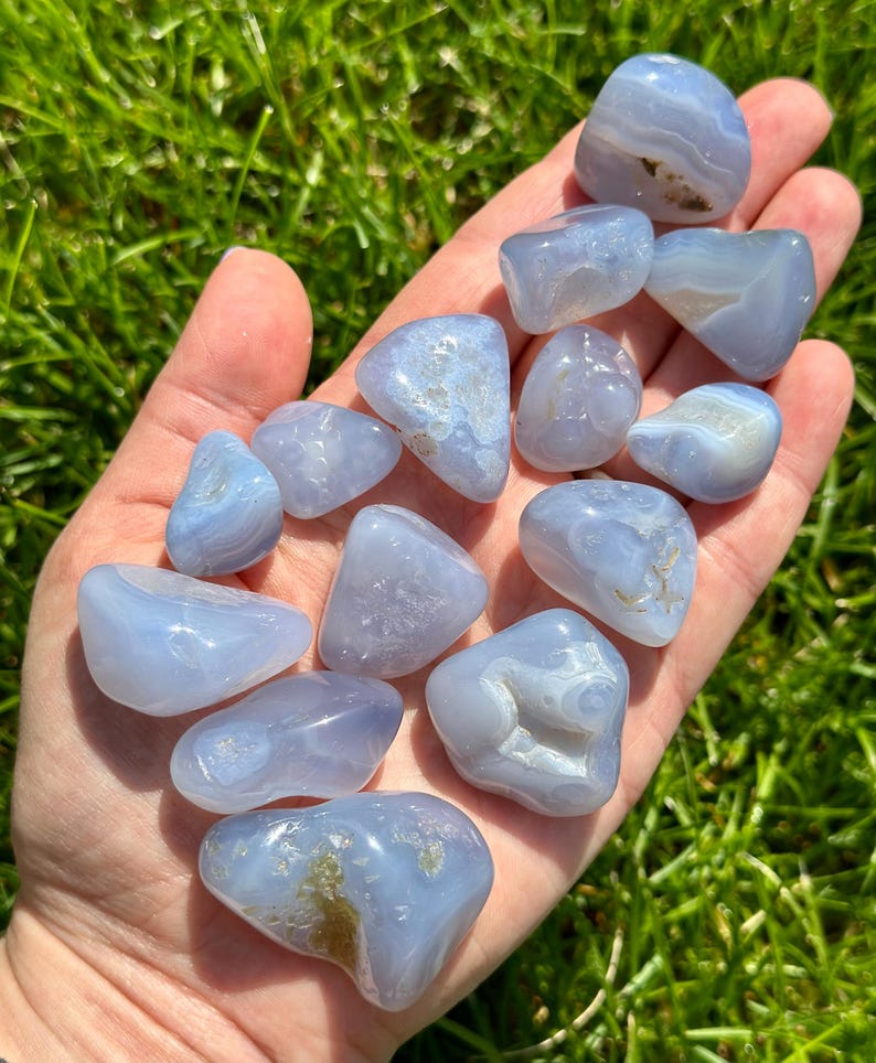 May include: A collection of smooth, tumbled blue lace agate stones. The stones vary in size and shape, with a pale blue color and white and brown inclusions. The stones are held in a hand against a backdrop of green grass.