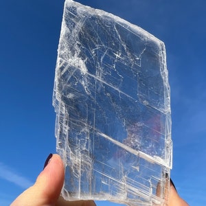 Puede incluir: Una gran losa de cristal de selenita transparente con capas y estrías visibles. El cristal se sostiene en una mano contra un fondo de cielo azul.