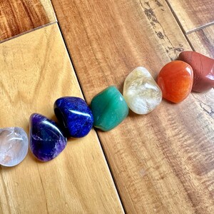 May include: Seven smooth, polished stones in a rainbow of colors, including clear, amethyst, blue, green, yellow, orange, and red. The stones are arranged in a line on a wooden surface.