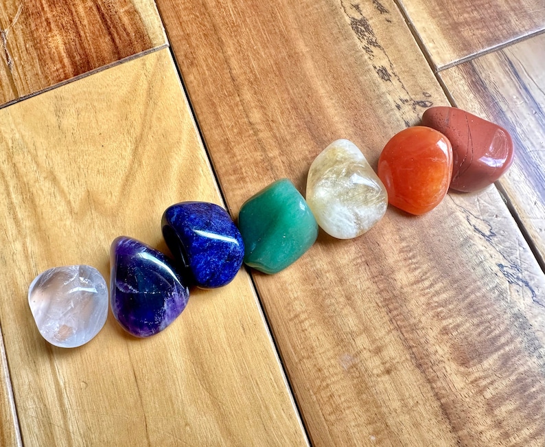 May include: Seven smooth, polished stones in a rainbow of colors, including clear, amethyst, blue, green, yellow, orange, and red. The stones are arranged in a line on a wooden surface.