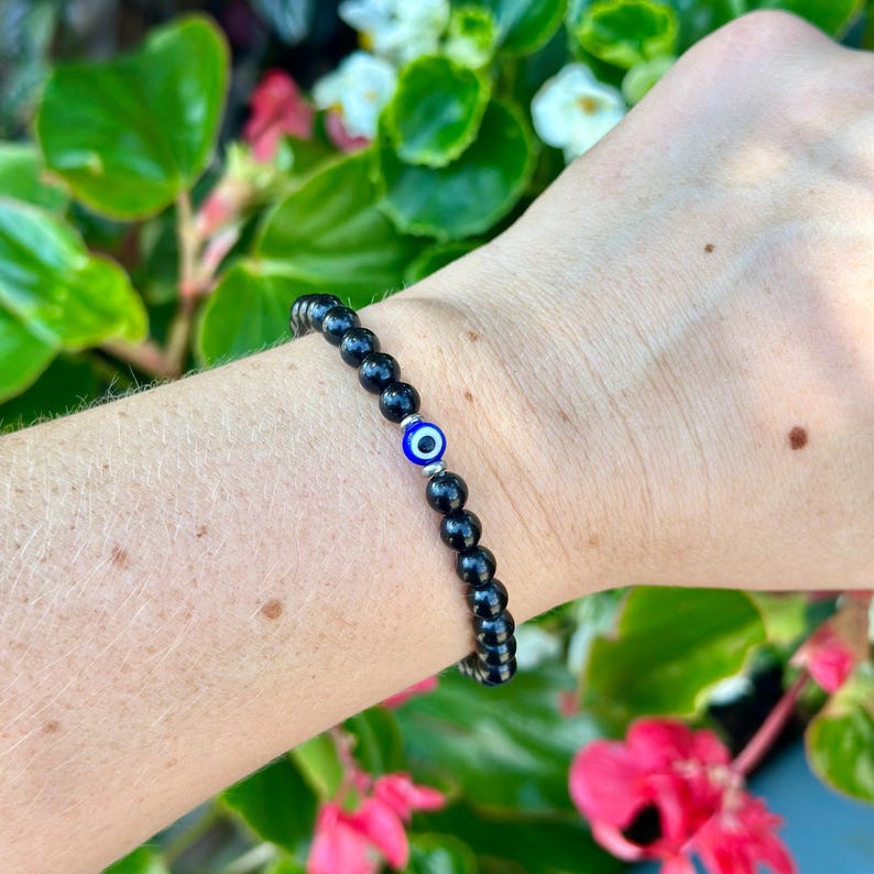May include: Black beaded bracelet with a blue and white evil eye charm. The bracelet is made of small, round, black beads and is worn on the wrist. The evil eye charm is a small, round bead with a blue center and white and black details.