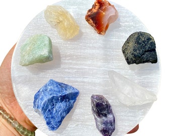 May include: A white selenite charging plate with seven different colored crystals arranged in a circle. The crystals are amethyst, clear quartz, carnelian, green aventurine, lapis lazuli, black obsidian, and citrine.