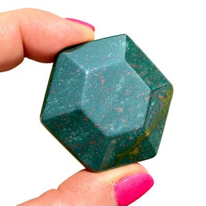 May include: A faceted, dark green gemstone with red speckles is held between fingers with pink nail polish. The gemstone has a geometric shape with multiple sides and a polished surface. The stone is a deep, rich green color.