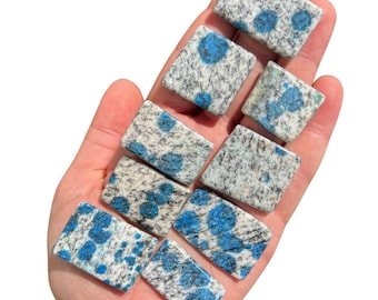 May include: A collection of eight polished blue and white speckled stone rectangles. The stones are arranged in a pyramid shape on a hand.