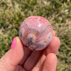 May include: A pink and white sphere with black and gray banding. The sphere is made of a polished stone with a smooth surface.