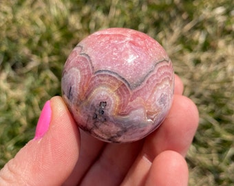 May include: A pink and white sphere with black and gray banding. The sphere is made of a polished stone with a smooth surface.