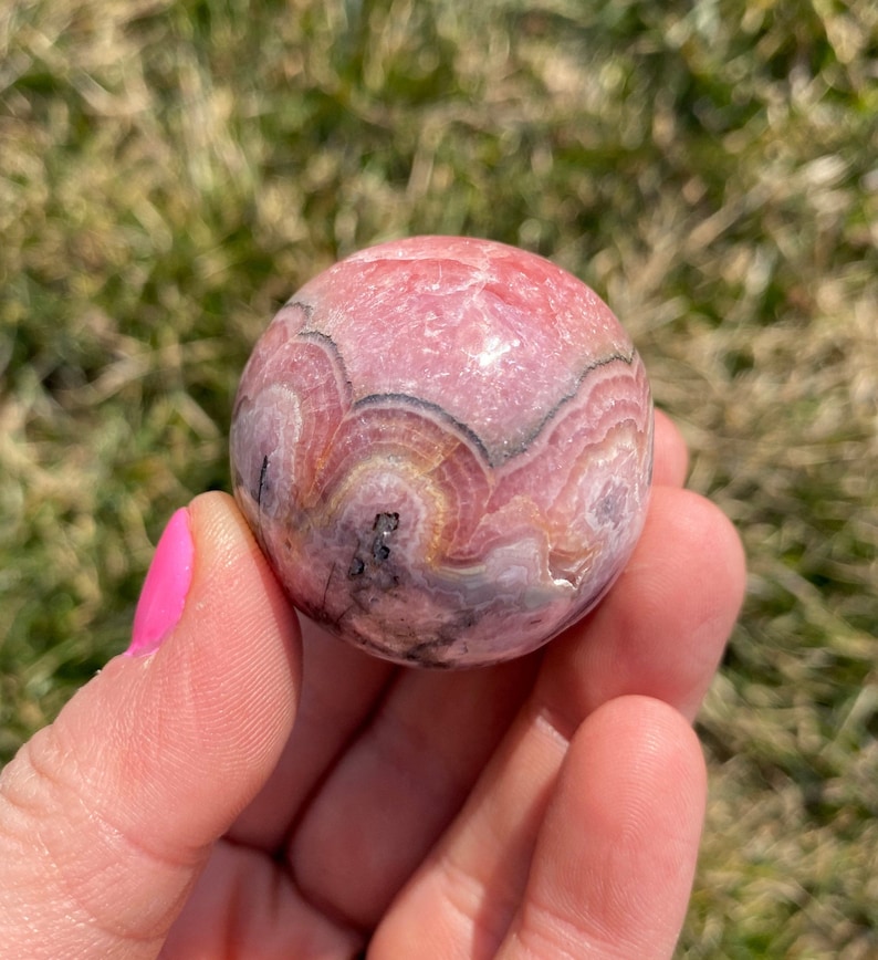 May include: A pink and white sphere with black and gray banding. The sphere is made of a polished stone with a smooth surface.