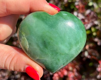 May include: A smooth, heart-shaped polished stone in a vibrant shade of green. The surface of the stone displays natural variations in color, including lighter and darker green tones, with a glossy finish that reflects light across its curved form.