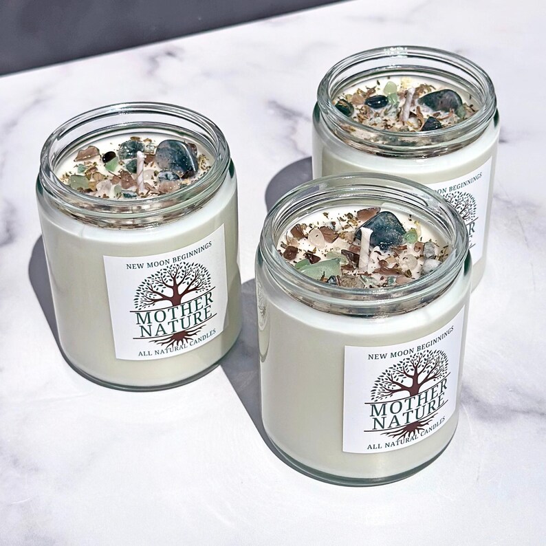 May include: Three glass candle jars with white wax and green and brown crystals inside. The jars have labels that say "New Moon Beginnings" and "Mother Nature All Natural Candles".