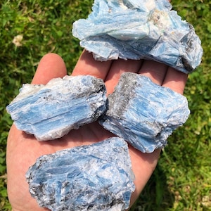 May include: Four rough, blue kyanite crystal specimens with a layered, striated texture. The crystals are a light blue color with some darker blue and black streaks.