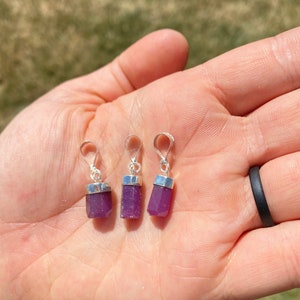 May include: Three small purple gemstone pendants with silver settings. The pendants are in the shape of a rectangular prism and are hanging from silver loops.