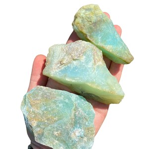 Puede incluir: Tres piedras de aguamarina en bruto de diferentes tama&ntilde;os, sostenidas en una mano. Las piedras muestran un color azul verdoso claro con &aacute;reas de color beige y marr&oacute;n. El fondo es blanco liso, resaltando la belleza natural de las piedras.